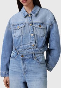 Chaqueta de mezclilla azul clara con un diseño recortado, grandes bolsillos frontales, botones en tono dorado y detalles de desgaste visibles. Textura suave y ajuste relajado.