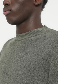 Κοντινό πλάνο του λαιμού και του κάτω μέρους του προσώπου ενός ατόμου που φοράει γκρι πουλόβερ με υφή και στρογγυλή λαιμόκοψη, πάνω σε απλό φόντο.