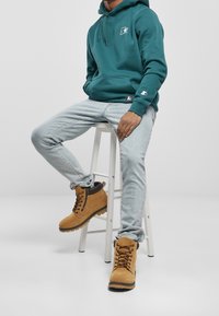 Sudadera con capucha color verde azulado con bolsillo tipo canguro y logo; jeans azul claro; botas marrones con cordones, detalles negros y cremallera metálica. Sentado en un taburete.