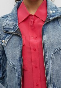 Jeansjacke mit weitem Kragen, hellblauer Farbe und silbernen Reißverschlüssen, über einem knopfärmeligen, leuchtend pinken Hemd mit passenden Knöpfen.