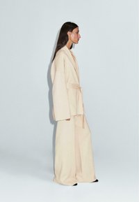 Jasnobeżowy, oversize'owy płaszcz z mieszanki wełny z szerokimi rękawami i paskiem, zestawiony z dopasowanymi spodniami o szerokich nogawkach. Gładka faktura i minimalistyczny design.