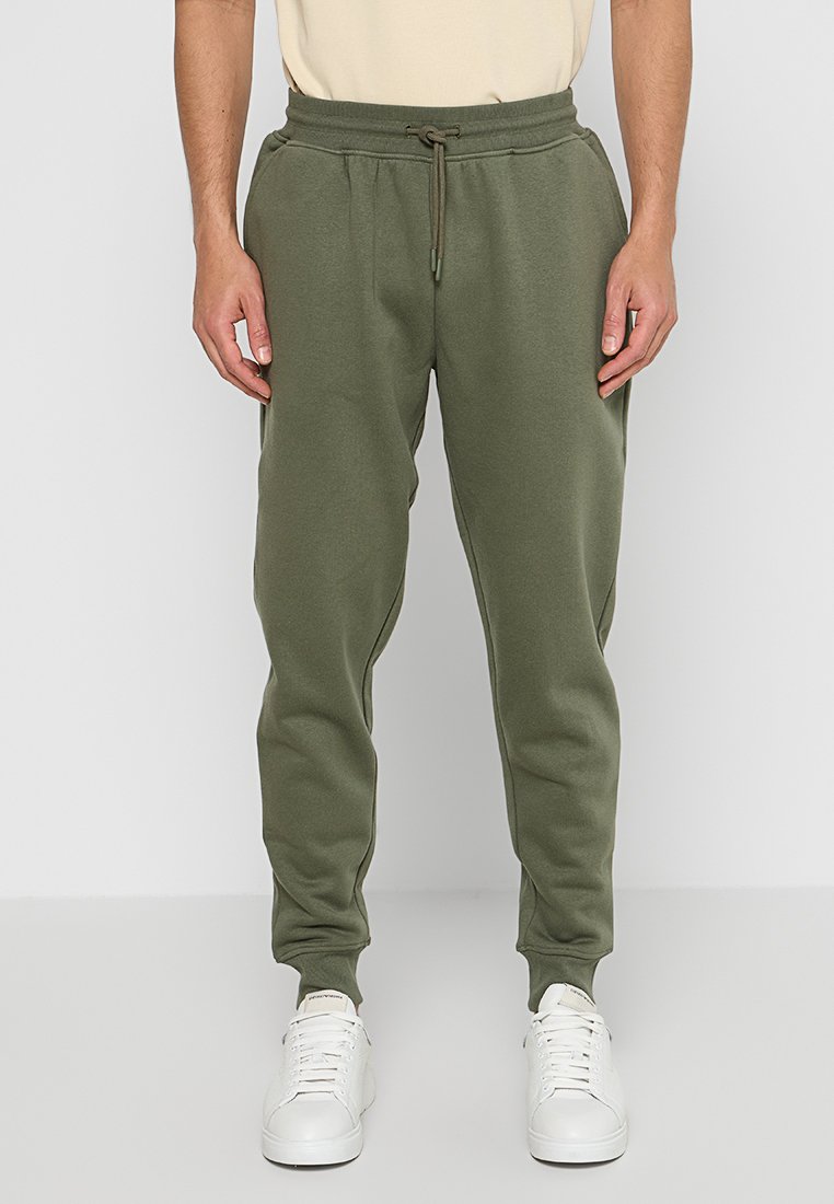 Pantaloni sportivi verdi con vita elasticizzata e cordoncino, vestibilità affusolata, tasche laterali e polsini a coste, indossati con sneakers bianche.