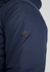 Giacca blu navy con tessuto liscio, una toppa nera testurizzata con testo in rilievo sul braccio sinistro e un design con cappuccio.