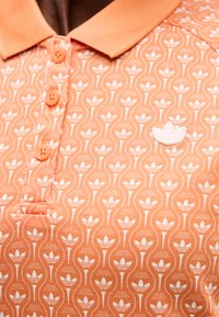 Πορτοκαλί πόλο μπλούζα με επαναλαμβανόμενο λευκό φυτικό μοτίβο τριφυλλιού και πατιλέτα με τρία κουμπιά. Λευκό κεντημένο λογότυπο τριφυλλιού στο στήθος.