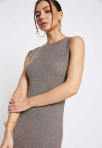 Rebrasta, brezrokavna obleka iz mehkega sivega blaga, ki se ponaša z oprijetim silhuetom in okroglim izrezom. Model prikazuje eleganten zlat nakit.