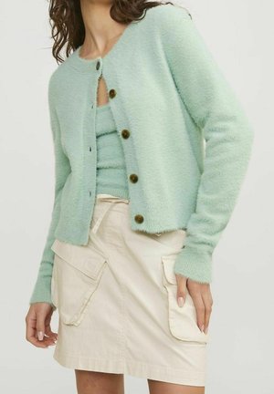 Femme portant un cardigan duveteux vert clair avec des boutons foncés sur un haut assorti, associé à une jupe beige avec de grandes poches avant.