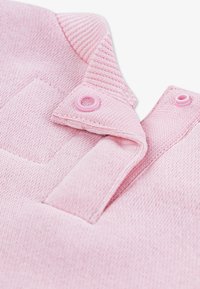 Hellrosa Polohemd mit einem gerippten Kragen, Kunststoffknöpfen und einem strukturierten Strickstoff. Auf der Vorderseite befindet sich ein quadratisches Aufnäher.