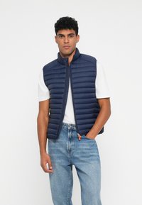 Marineblå pufvest med vandret quiltning, frontlynlås og orange lynlåsdrager. Sættes sammen med en hvid T-shirt og lyseblå jeans.