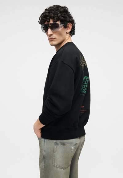 Άνδρας με σγουρά μαλλιά που φοράει μεγάλα γυαλιά ηλίου με διάφανο πλαίσιο, μαύρο φούτερ με πολύχρωμο σχέδιο στην πλάτη και ξεθωριασμένο γκρι τζιν, με τα χέρια στις τσέπες.