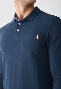 Mornarska polo majica dugih rukava od teksturiranog materijala, s trodijelnim kopčanjem i džepom na lijevoj strani prsnog koša s crvenim, bijelim i plavim detaljem.