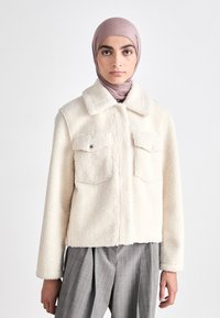 Cremefarbene Kunstfelljacke mit Kragen, zwei Brusttaschen und silbernen Knöpfen. Kombiniert mit grau gestreiften Hosen.