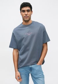 Γκρι βαμβακερό t-shirt με κοντά μανίκια και στρογγυλή λαιμόκοψη. Διαθέτει λογότυπο "Calvin Klein JEANS" σε ροζ στο μπροστινό μέρος. Συνδυάζεται με ανοιχτόχρωμο μπλε τζιν.