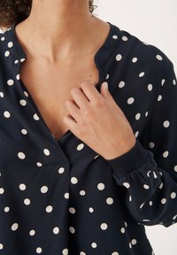 Main d'une femme ajustant le col d'un chemisier bleu marine à pois blancs, avec un col en V à encoche et des poignets côtelés.