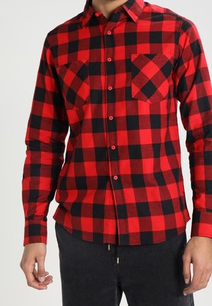 Uomo che indossa una camicia a quadri rossa e nera a maniche lunghe con due tasche sul petto, abbinata a pantaloni neri.