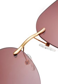 Kulaté sluneční brýle se gradientními burgundskými skly, zlatým kovovým mostem a průhlednými nosními podložkami. Mají elegantní, minimalistický design.