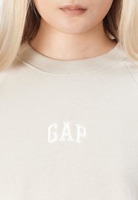 Primer plano de una persona que lleva un sudadera beige con el logotipo bordado en blanco "GAP" centrado debajo del escote.