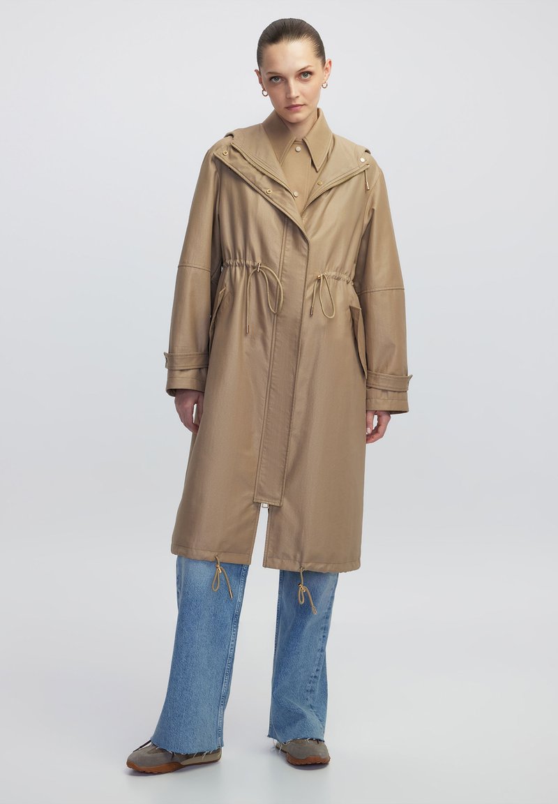 Trench coat beige leggero con vita regolabile, colletto ampio e tasche, abbinato a jeans blu a gamba larga e calzature marroni.