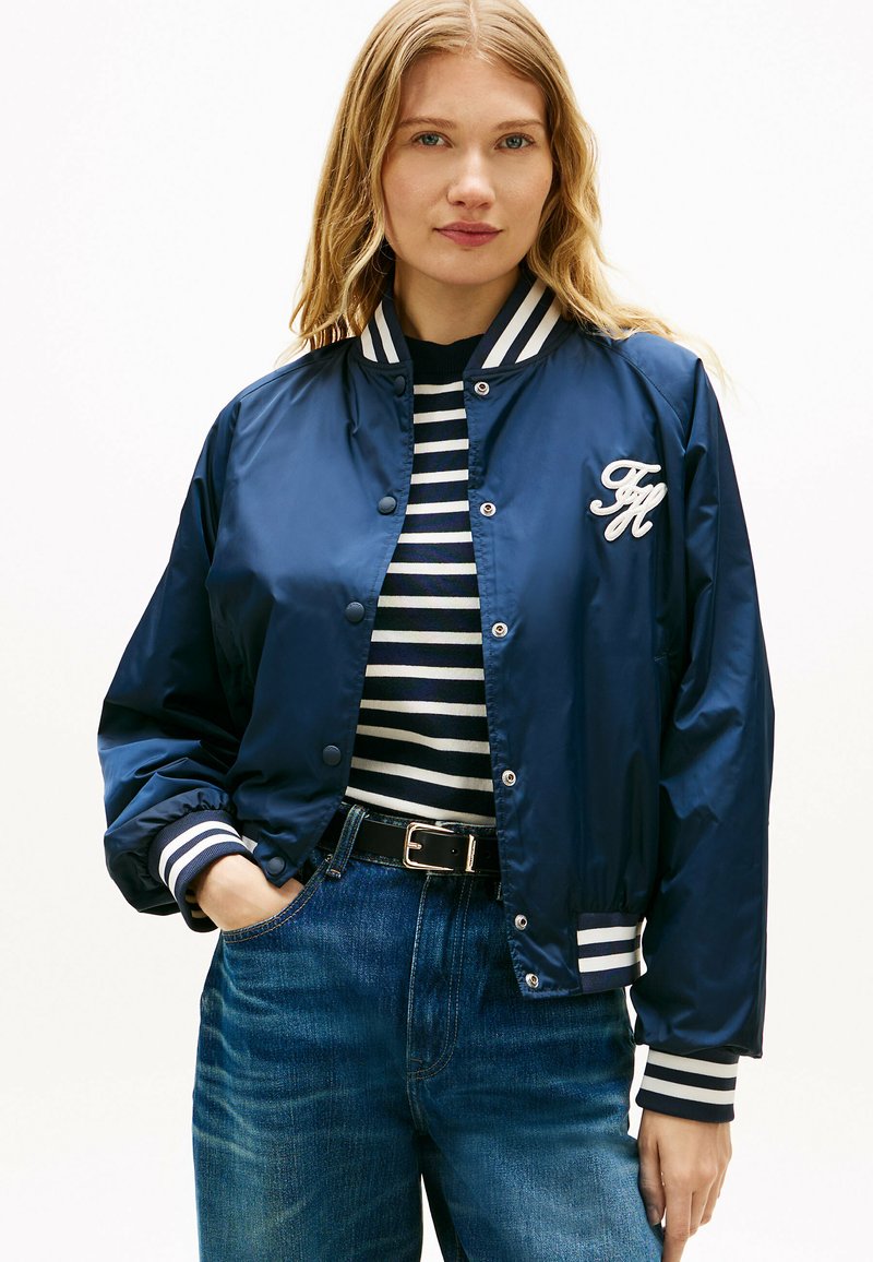 Woman wearing a navy blue bomber jacket with white striped cuffs and collar, over a black and white striped shirt and blue jeans.