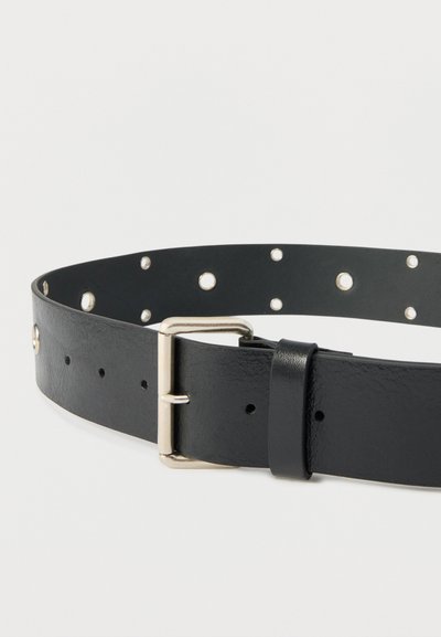 Ceinture en cuir noir avec boucle argentée et plusieurs œillets métalliques ronds, sur fond blanc lisse.