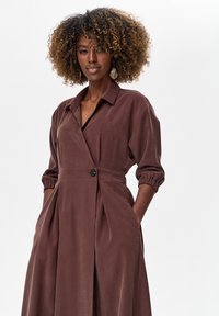 Brązowa sukienka z długim rękawem, z kołnierzykiem, taliowana w pasie i z plisowanym dołem. Cechy charakterystyczne to czarny guzik i fakturowana tkanina.