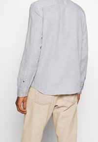 Camicia a maniche lunghe grigio chiaro con tessuto strutturato, polsini con bottoni e orlo arrotondato, abbinata a pantaloni beige.