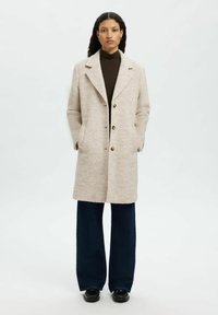 Beige Wollmischungsjacke mit einem Reverskragen, frontalen Knöpfen und seitlichen Taschen, kombiniert mit dunklen weitgeschnittenen Hosen und schwarzen Schuhen.