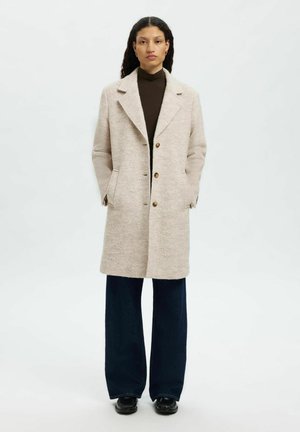 Manteau en mélange de laine beige avec un col cranté, des boutons sur le devant et des poches latérales, associé à un pantalon large noir et des chaussures noires.