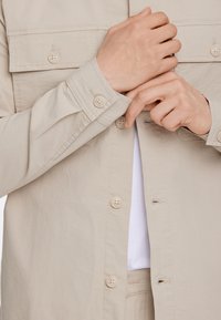 Beige button-up skjorta med en texturerad tyg, med två bröstfickor och standardknappar. En vit skjorta syns under.