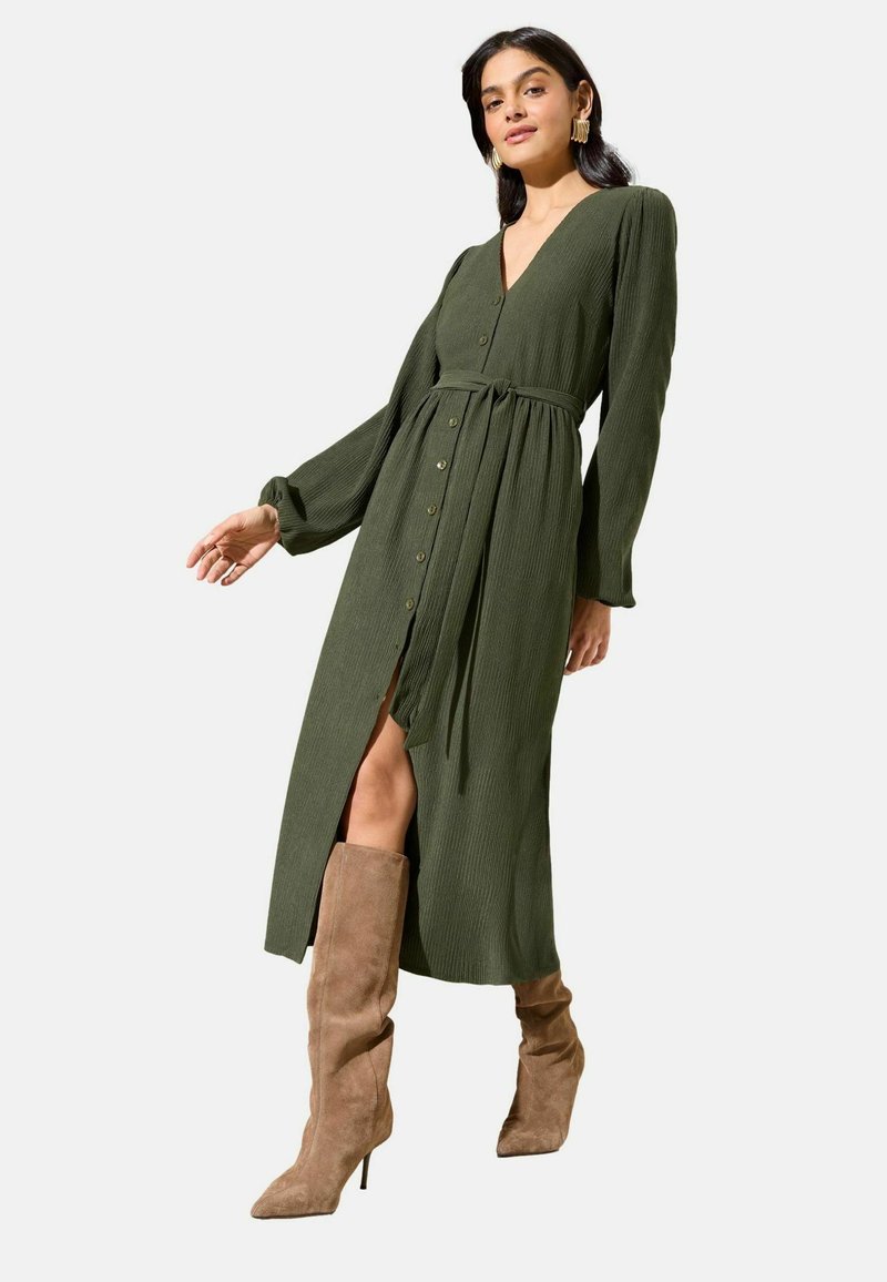 Olijfgroene jurk met lange mouwen, V-hals, knopen aan de voorkant en tailleband. Geribbelde structuur en een voorste split. Gecombineerd met beige laarzen tot aan de knie.