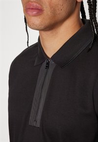Polo noir avec fermeture éclair, dotée d'un panneau de tissu contrastant au niveau du col, de manches courtes et d'une texture lisse.