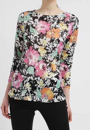 Mujer lleva una blusa de manga larga con estampado floral en rosa, naranja, amarillo y verde sobre un fondo blanco y negro combinada con pantalones negros.