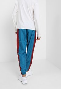 En person i blå träningsbyxor med röda sidsnören, vita sneakers och en långärmad vit skjorta, står i ett vitt rum.