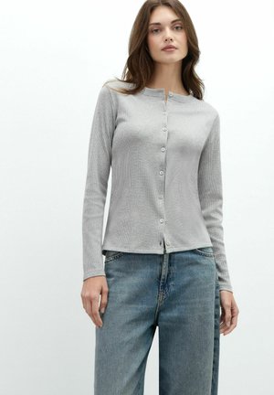 Femme portant un cardigan côtelé gris clair boutonné et un jean bleu ample, debout devant un fond blanc.