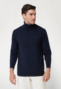 Maglione dolcevita blu navy con colletto e polsini a coste, realizzato in un tessuto morbido. Presenta un piccolo logo sul petto. Indossato con pantaloni chiari.