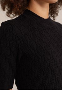 Kvinna som bär en svart kortärmad tröja med kabelstickning och en ribbad hög hals, som visar ett texturerat stickat mönster och lockigt hår.