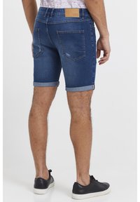 Mörkblå denimshorts med uppvikta fållar, fem fickor och en brun läderetikett på midjebandet. Bärs med svarta sneakers.