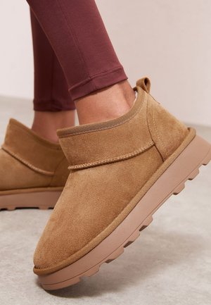 Personne portant des bottines en daim beige à enfiler avec des semelles épaisses en caoutchouc et des leggings bordeaux sur un sol clair.
