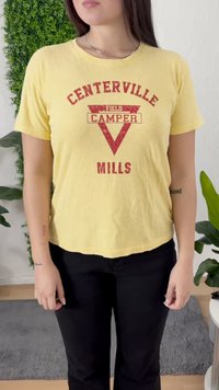Žuta majica s kratkim rukavima sa izblijedjelim crvenim grafikom koja prikazuje natpis "CENTERVILLE CAMPER" unutar trokuta, uparena s crnim hlačama.