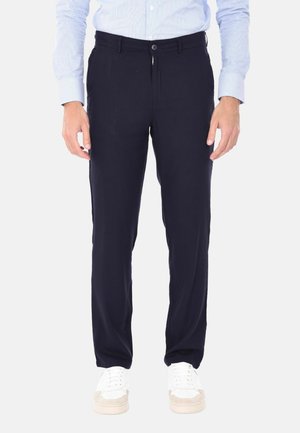 Uomo con pantaloni neri a gamba dritta, camicia azzurro chiaro abbottonata e sneakers bianche, in piedi contro uno sfondo chiaro e semplice.