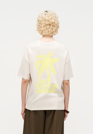 Persoon met kort krullend blond haar die een lichtbeige T-shirt draagt met een gele palmboom en de tekst 'DE ZON MAAKT JE BLIJ' op de achterkant.