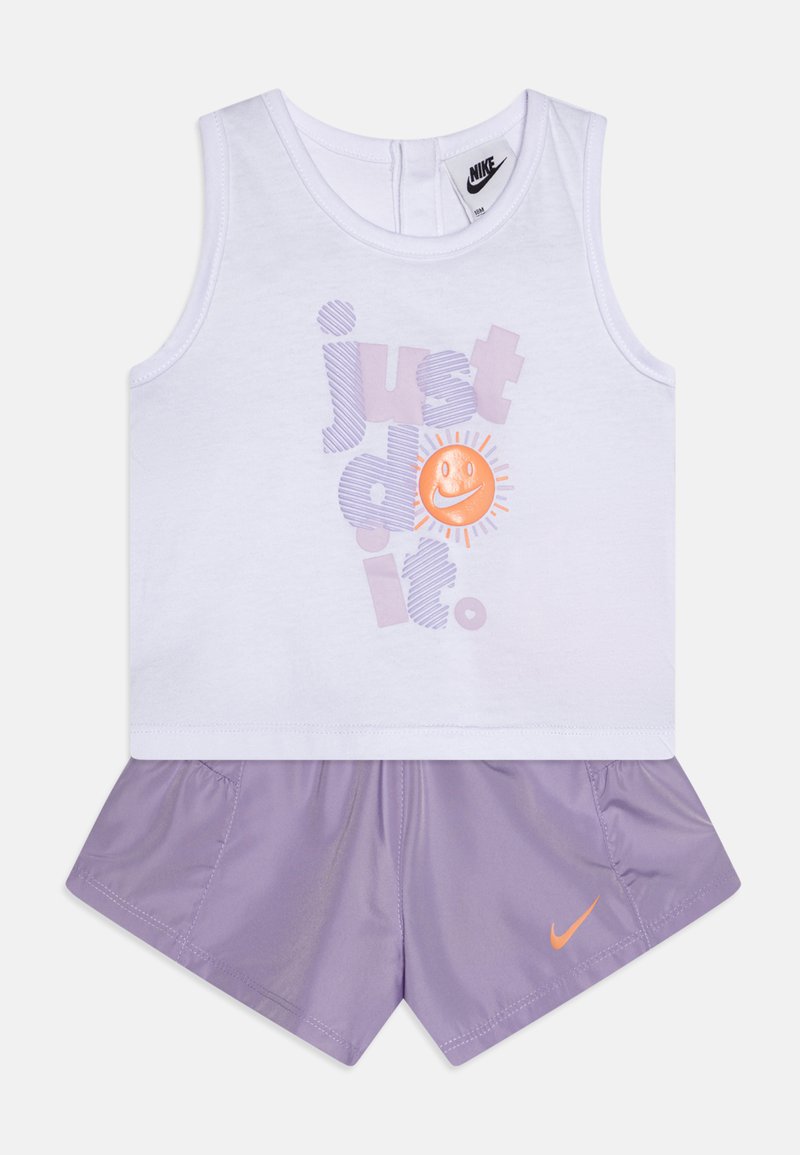 Bela brezrokavna majica z napisom "samo naredi to" in nasmejanim sončnim grafikom. V kompletu z lila kratkimi hlačami, ki imajo oranžni Nike logotip.