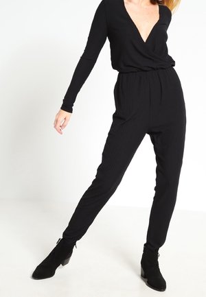 Czarny prążkowany kombinezon z głębokim dekoltem w serek i długimi rękawami, z elastycznym pasem i zwężanymi nogawkami, zestawiony z czarnymi botkami za kostkę.