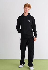 Sudadera negra con un logo en la parte frontal, combinada con pantalones negros. El modelo lleva zapatillas blancas, con un fondo neutro.