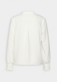 Blouse blanche à manches longues avec poignets froncés et un pli dans le dos, présentée sur un fond clair uni.
