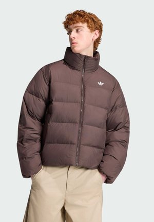 Junge Person trägt eine braune Steppjacke mit hohem Kragen und beige Hose, sieht zur Seite und hat die Hände in den Taschen.