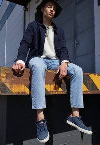 Marinblå mockaskor med vita sulor; modellen sitter på en rostig metallkant iförd en mörk jacka och ljusa jeans, med en bredbrättad hatt.