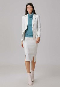 Femme portant un blazer blanc et une jupe, un pull à col roulé teal, et des bottes blanches à la cheville, se tenant devant un fond gris uni.