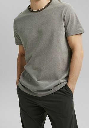 Homme portant un t-shirt gris à manches courtes décontracté et un pantalon noir, debout les mains dans les poches devant un fond uni.