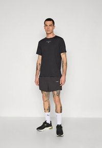 T-shirt negra de manga curta com logótipo, combinada com calções escuros. Modelo usando sapatilhas pretas desportivas e meias brancas até ao tornozelo. Design simples e desportivo.