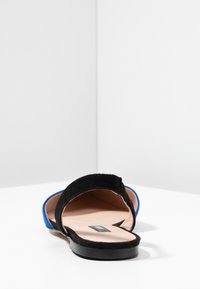 Wildledersandale mit schwarzem Obermaterial und blauen Akzenten, offenes Zehendesign und flache schwarze Sohle. Das Innere besitzt eine weiche beige Fütterung.